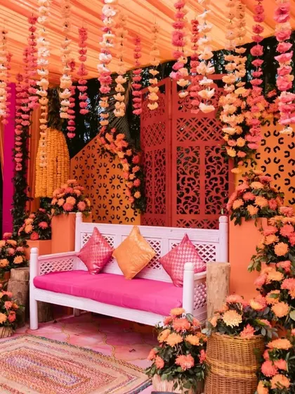 A cozy and vibrant photo corner at a mehendi. A white bench with pink cushions is surrounded by orange floral arrangements and a patterned backdrop, perfect for guest photos.