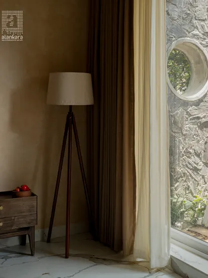 A quiet corner in a well-designed bedroom. This image highlights the interplay of textures, from the soft, layered curtains to the stone wall visible through the circular window and the classic wooden tripod floor lamp.