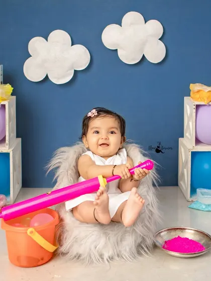 This happy baby is ready for Holi with her pichkari and bucket, sitting in a fluffy chair.