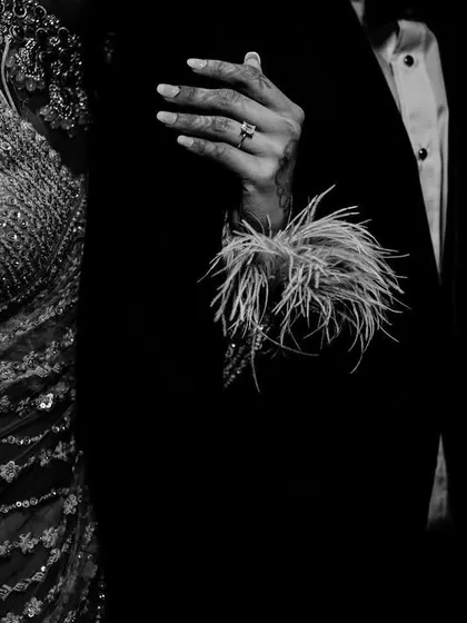 A detailed black and white shot of the couple's hands and attire, focusing on the textures of the bride's sequined dress and feathered sleeves.