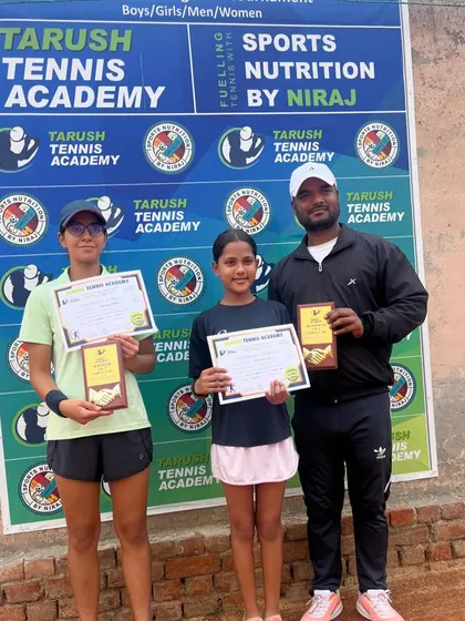 Eshanya Khattar and another player pose with their coach and awards at the Tarush Tennis Academy AITA event, in front of the official tournament backdrop.