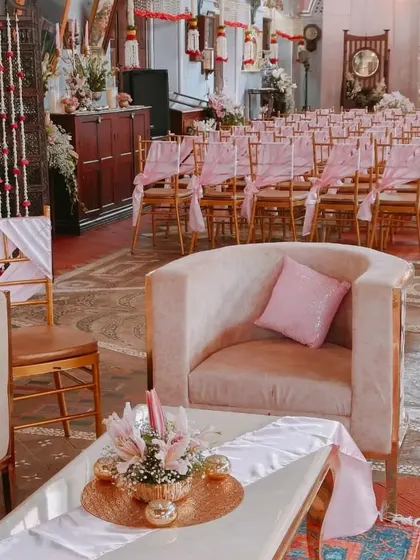 A view of an indoor wedding ceremony setup, highlighting the comfortable lounge seating in the foreground and the elegantly decorated guest chairs with pink sashes in the background.