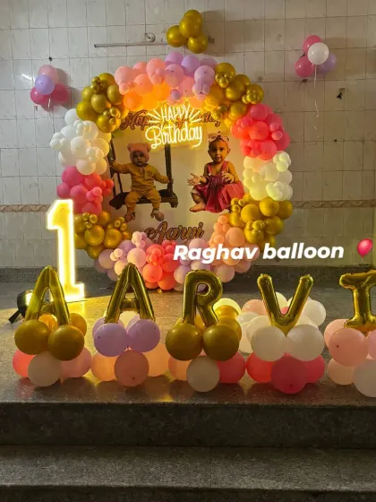 A personalized first birthday decoration with the baby's name "AARVI" in foil balloons. The setup includes a circular balloon arch with a custom photo backdrop and a light-up number '1'.