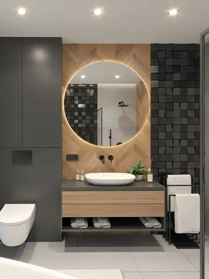 This modern bathroom design uses a bold, 3D black tile accent wall behind a backlit round mirror. The floating wood vanity and wall-mounted toilet keep the floor clear, making the space feel larger.
