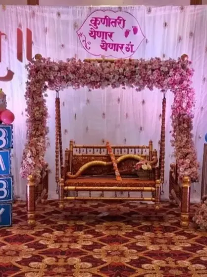 A fun and colorful baby shower decoration featuring large, custom-made cutouts of a pink and a blue baby pram. The central jhula is framed with flowers, and a Marathi sign "Kuniतरी येणार येणार गं" adds a personal, local touch to the celebration.