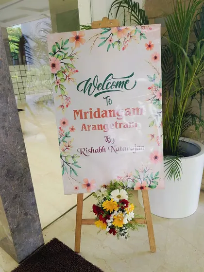 A simple and elegant welcome board for a Mridangam Arangetram. The floral-printed sign is placed on a wooden easel and decorated with a fresh flower arrangement.