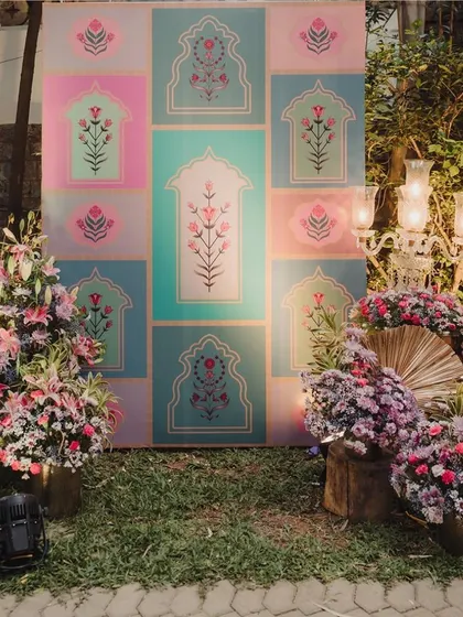 A beautiful photo backdrop featuring pastel-colored panels with traditional Indian motifs. The setup is flanked by lush floral arrangements, creating a perfect blend of modern and traditional design.