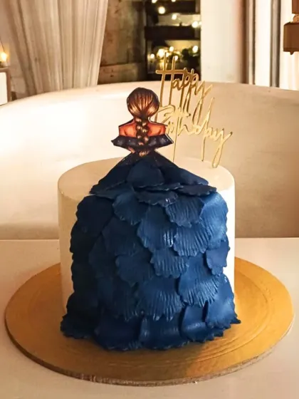 An artistic and fashionable cake featuring a topper of a girl in a flowing blue gown created with textured buttercream petals.
