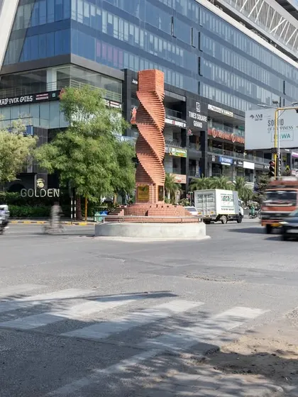The twisted brick column in its wider urban context, surrounded by modern commercial buildings. The design bridges the gap between Ahmedabad's traditional brick architecture and its contemporary growth.