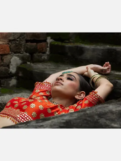 An artistic shot of a model in a traditional orange blouse, lying on ancient stone steps. The composition and soft lighting create a serene and timeless mood for this ethnic wear piece.