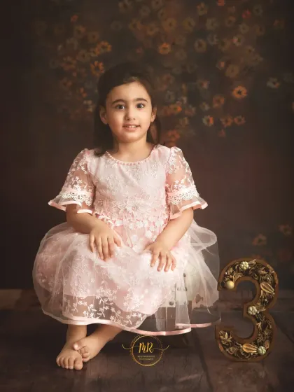A three-year-old girl in a lovely pink lace dress poses next to a decorative number "3".