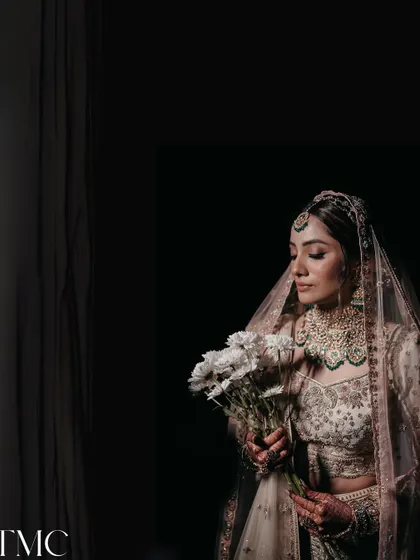 A dark and moody bridal portrait. The bride, holding a simple bouquet of white flowers, is illuminated by a single light source, creating a dramatic, high-contrast image that is both timeless and modern.