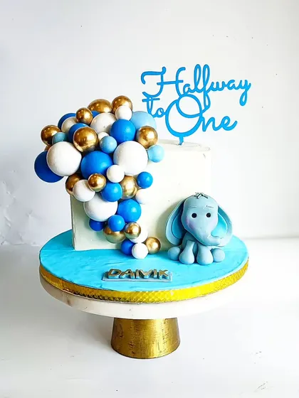 A cute half-birthday cake to celebrate being 'Halfway to One'. This design features a baby elephant and a blue and gold balloon cascade.