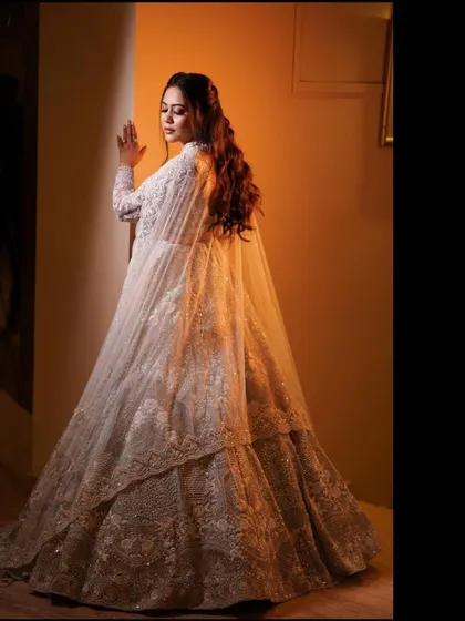 An elegant, full-length portrait of the bride in her white caped lehenga. The warm lighting and her pose create a look of regal sophistication and modern bridal style.