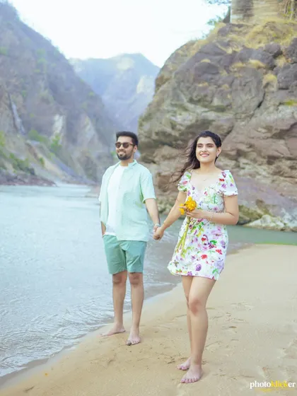 A couple walks hand-in-hand along the water's edge in Rishikesh, with the woman holding a bouquet of sunflowers, adding a pop of color.
