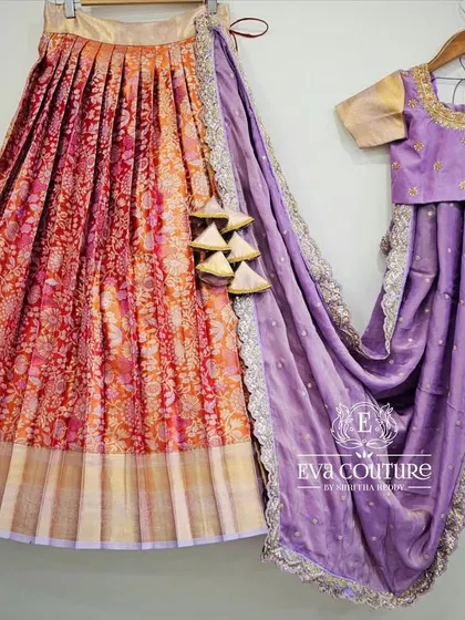 A beautiful red and orange Banarasi lehenga with a contrasting lilac half-saree style dupatta. The lilac blouse features delicate gold maggam work around the neckline.