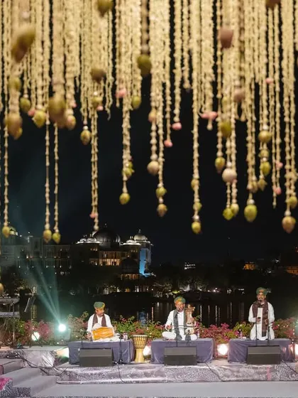A magical Sangeet night in Udaipur with musicians performing under a ceiling of suspended tuberoses and marigolds, with the shimmering lake and palace in the background.