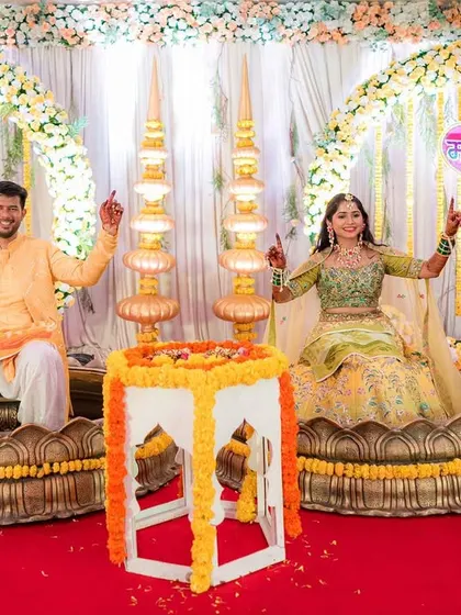 The happy couple at their Haldi ceremony. The backdrop features large floral rings and traditional brass lamps, perfectly complementing the joyous occasion.
