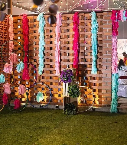A creative photo corner at a Mehendi event, featuring colorful tassels and decorative bicycle wheels against a brick wall. This provides a fun and quirky spot for guest photos.