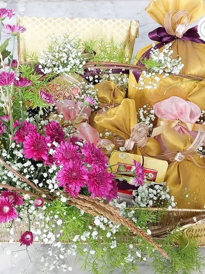 A luxurious hamper overflowing with fresh magenta flowers, baby's breath, and beautifully wrapped traditional gifts and dry fruits in gold and pink potlis.