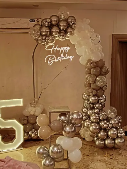 A chic 25th birthday decoration with a modern balloon arch. The setup uses a mix of silver, nude, and white balloons with a neon sign and large marquee numbers.