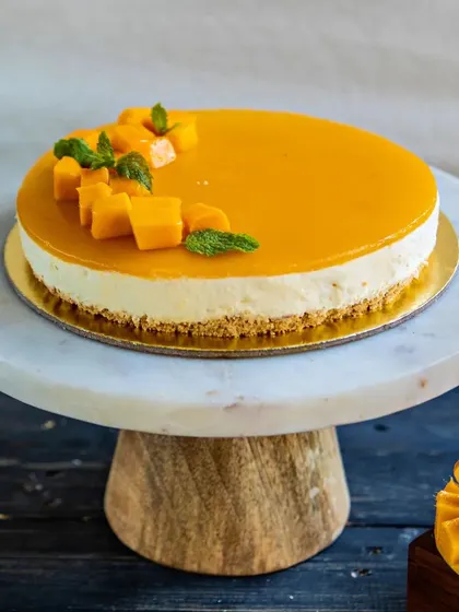 Our Mango Cheesecake, elegantly presented on a marble stand. It features a smooth mango glaze and is topped with fresh mango chunks and mint, making it a refreshing seasonal dessert.