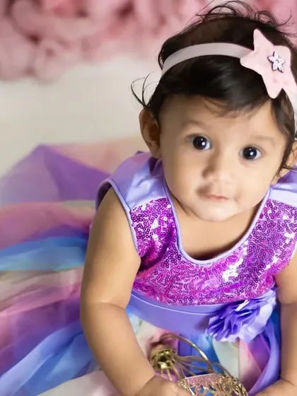 A close-up of a beautiful baby girl in a rainbow dress, looking right at the camera with her big, expressive eyes.