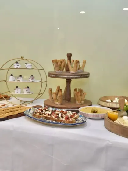A full view of a sophisticated grazing table setup for a clinic launch. The clean, white tablecloth provides a perfect canvas for tiered platters of canapés, bruschetta, and a central charcuterie board, all elegantly arranged with custom menus.