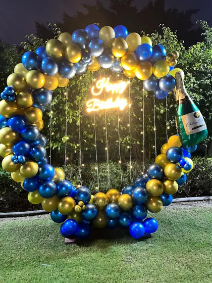 Perfect for an adult celebration, this outdoor setup features a circular balloon arch in royal blue and gold. The design is completed with a champagne bottle balloon, a central neon sign, and delicate fairy lights.
