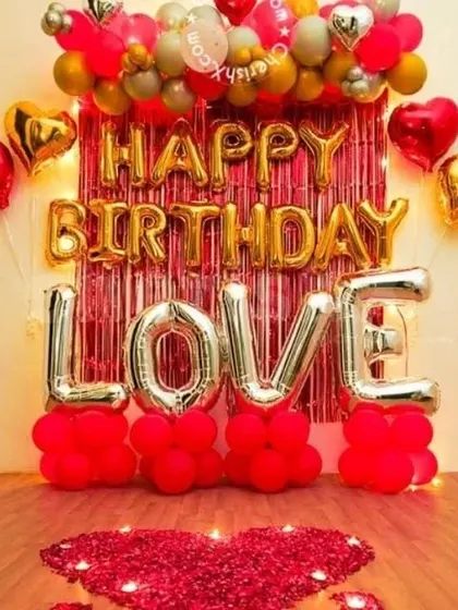 A vibrant romantic birthday decoration with "LOVE" letter balloons, a heart made of rose petals, and a backdrop of red and gold balloons.