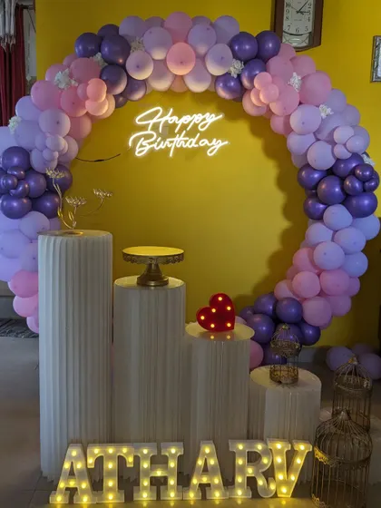 A stylish birthday setup with a circular balloon arch in shades of purple and pink. The decor is completed with a 'Happy Birthday' neon sign and modern white plinths.