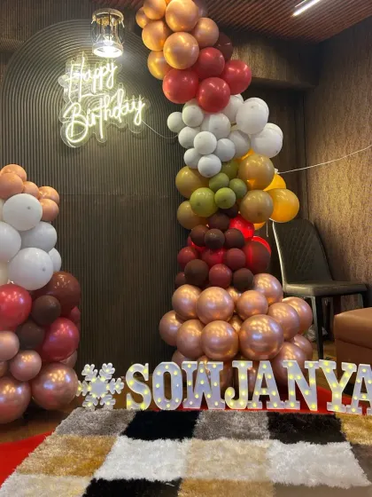 A cozy and stylish birthday corner. The decor features a unique balloon garland in earthy tones against a dark, textured wall, with a 'Happy Birthday' neon sign and marquee letters.