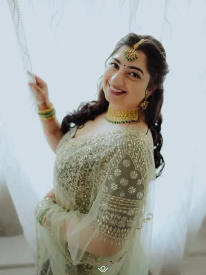 A happy bride in a light green sequined saree, her bright smile captured against a soft, backlit curtain.