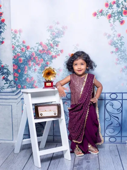 A stylish little girl posing confidently in a modern purple saree. The vintage props like the radio and gramophone add a classic touch to this fashionable child portrait.