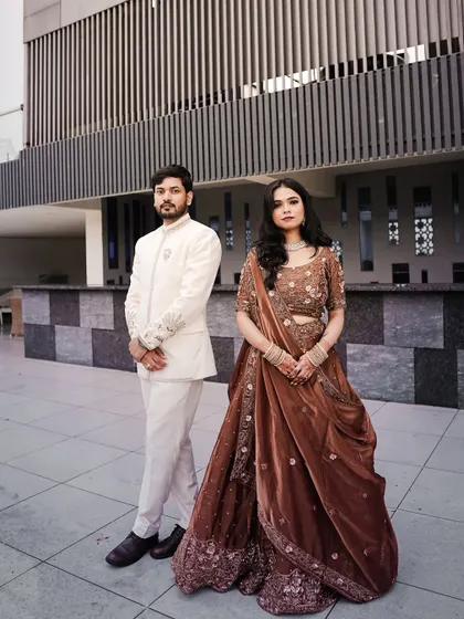 A full-length shot of Piyush and Anjali, showcasing their coordinated engagement outfits. The groom is in a sophisticated white sherwani, and the bride wears a beautiful brown embellished lehenga.