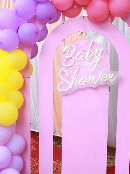 A close-up of a pink arch with a "Baby Shower" neon sign, decorated with a pastel rainbow balloon arrangement.