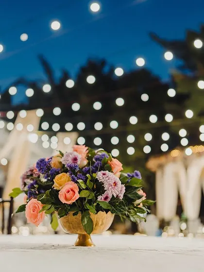 A colorful floral centerpiece on a guest table, with the beautifully lit Sangeet stage creating a lovely bokeh effect in the background.