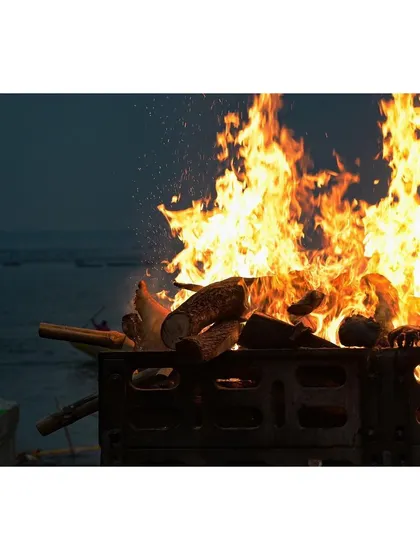 A close-up of a funeral pyre burning fiercely, with sparks flying against the dark backdrop of the river. This image isolates the element of fire, central to the Hindu ritual of cremation.