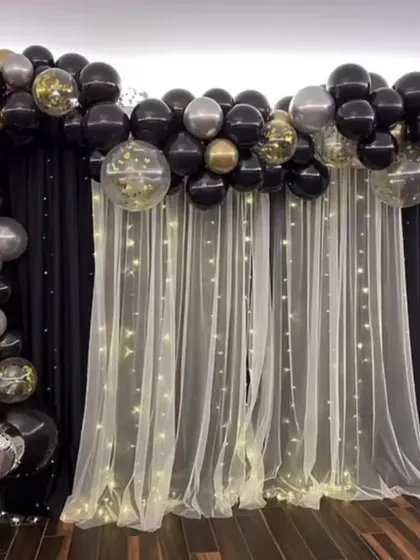 An elegant backdrop created with black and sheer white curtains and fairy lights. The look is framed by a black, gold, and silver balloon arch, making it suitable for any formal event.