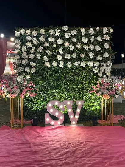 A personalized photo spot with the couple's initials in large, lit-up letters. This is set against a white floral wall and flanked by decorative light panels, perfect for outdoor evening events.