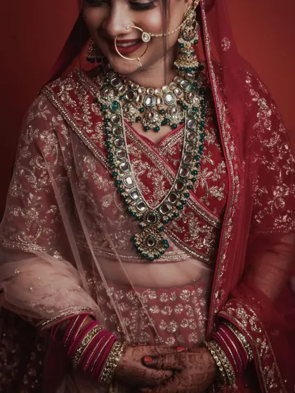 Capturing the complete bridal look, this image shows the bride in her magnificent red and gold lehenga. Her shy smile and the way she holds her hands convey a sense of gentle grace and excitement on her wedding day.