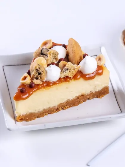 A slice of the Caramel Seasalt Cheesecake, showing off all the delicious toppings. It's a party of textures with cookie chunks, hazelnuts, and smooth caramel.