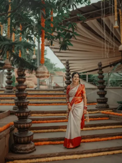 The bride stands elegantly on steps adorned with marigolds, her traditional white and red saree looking absolutely stunning.