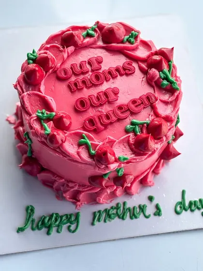 A special cake for Mother's Day with the message "Our Moms Our Queen." The vibrant pink buttercream is decorated with piped cherries for a fun and festive look.