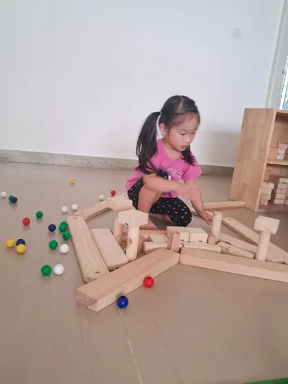 A child creates a long, winding path with wooden blocks and colorful marbles. This is a self-directed experiment in physics and design, as she figures out how to make the marbles roll.