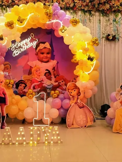 A fairytale first birthday celebration with a Disney Princess theme. This setup includes a circular backdrop with a photo, a warm-toned balloon arch, and cutouts of Cinderella, Snow White, and other beloved princesses.