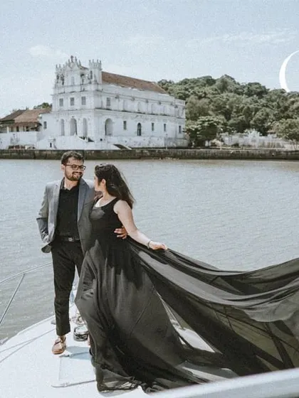 A cinematic pre-wedding shot on a yacht in Goa, with the historic Divar Island church in the background. The bride's long black dress adds a touch of drama and elegance.