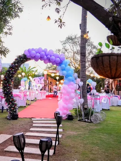 An outdoor birthday party entrance featuring a beautiful balloon arch in shades of purple, blue, and pink, leading guests down a red carpet.