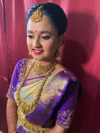 A happy bride moments before her ceremony. The off-white and purple silk saree is neatly pleated, and the simple, elegant updo is finished with a maang tikka, creating a classic and radiant bridal look.