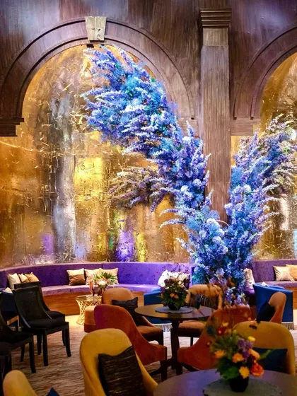 Dramatic plumes of blue-dyed florals rise against textured gold walls, creating a bold and artistic statement in the Sangeet lounge.